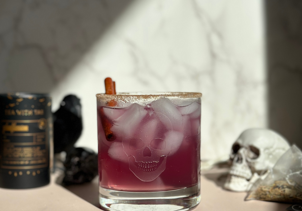 Glass of pink iced tea with ice cubes, cinnamon stick garnish, and skull design on the glass, beside a Tea With Tae tea tube and tea sachet.