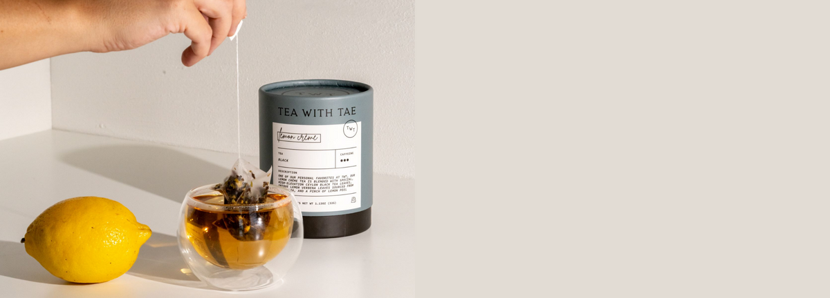 Hand lifting a tea bag from a glass cup of tea beside a Tea With Tae tea tube and whole lemon on a white surface.