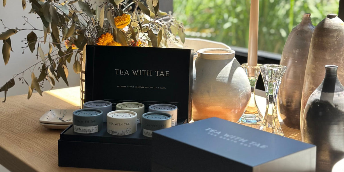 Open black Tea With Tae gift box displaying six mini tea tubes on a wooden table in natural sunlight, beside decorative vases and candles.