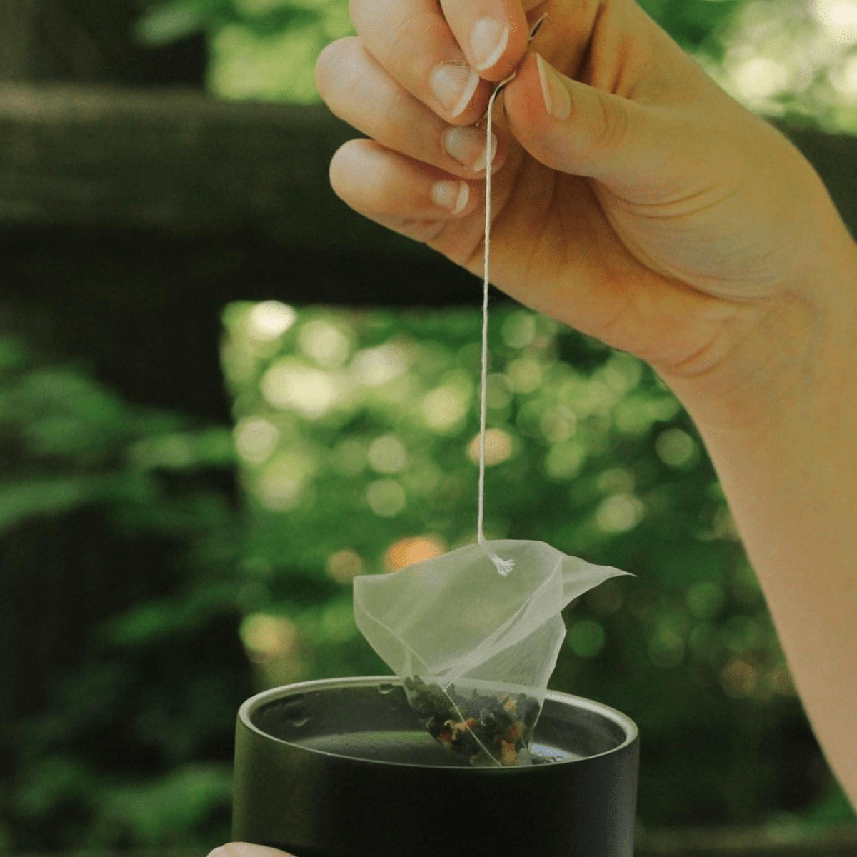 Hand lifting a tea bag from a black cup outdoors, with blurred green garden background.