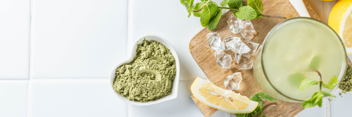 Top view of a green iced drink beside lemon slices, fresh mint, ice cubes, and green powder on a light surface.