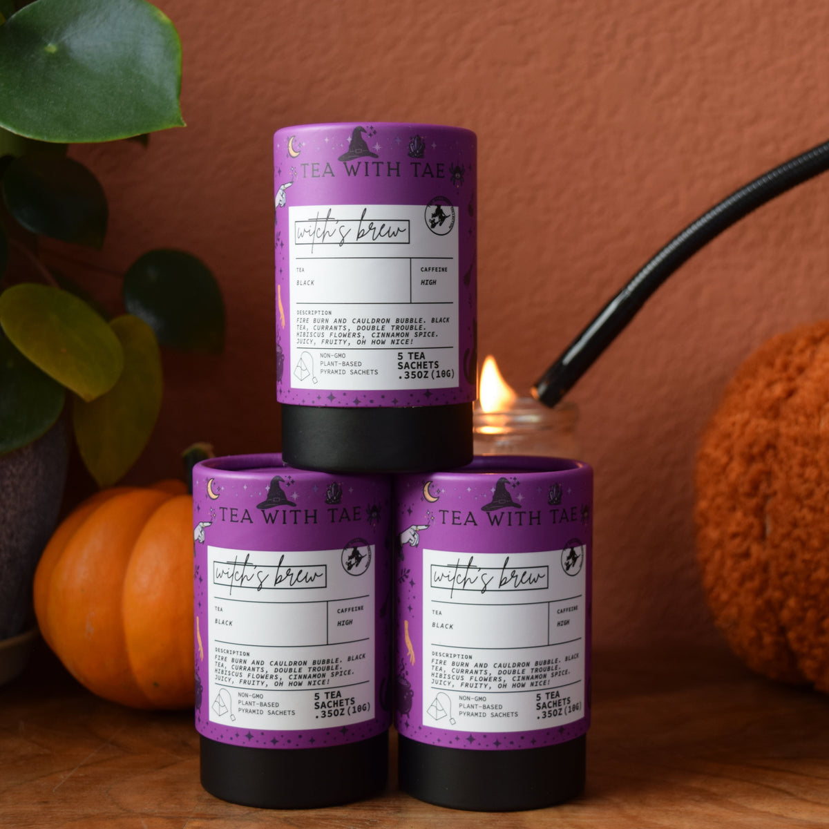 Three mini tea tubes with purple labels and black bases are arranged on a wooden surface, with one candle lit using a blue-handled torch. The scene includes decorative pumpkins, a potted plant, and a warm terracotta wall in the background.

