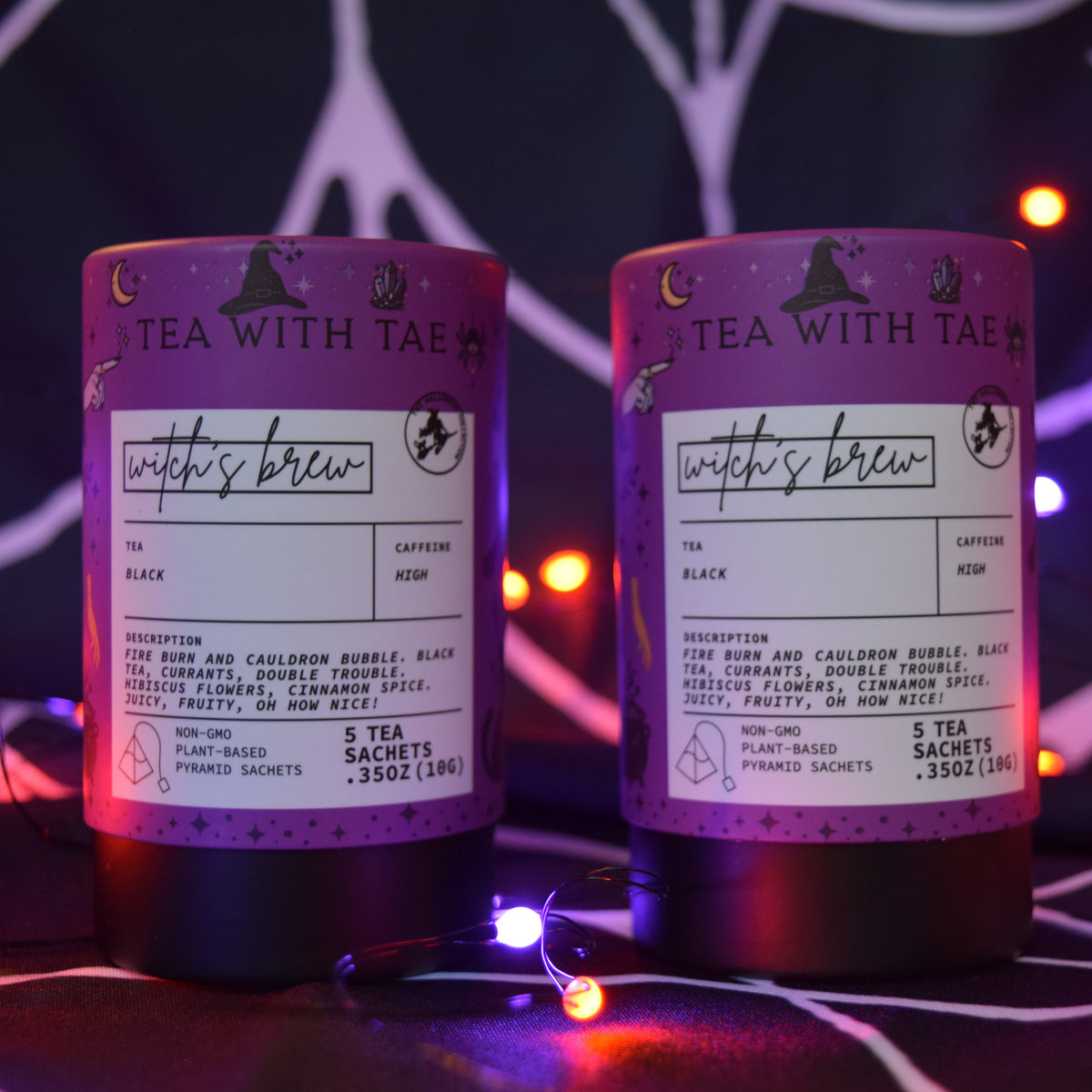 Two cylindrical tea product containers labeled 'witch's brew' and 'tea with tae' are shown on a dark surface with decorative fairy lights in the background.